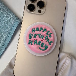 May include: A gold smartphone with a pink and green phone grip attached. The phone grip is round and says "Happee Birthdae Harry" in green letters. The phone has three camera lenses.