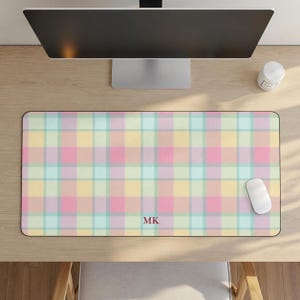 May include: A rectangular desk mat featuring a pastel plaid pattern in shades of yellow, pink, and light blue. The mat has a smooth surface and includes the letters MK printed in a dark red font centered near the bottom edge of the mat.