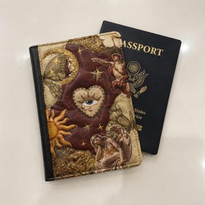 May include: A rectangular passport cover featuring a collage of a golden crescent moon, a sun, a central heart with an eye, and classical figures. The cover has a black spine and is displayed next to a dark blue United States passport.