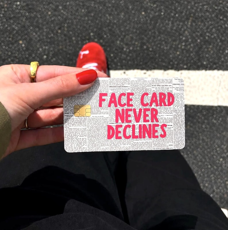 May include: A rectangular credit card skin featuring a black and white newspaper print background with the bold, bright pink text FACE CARD NEVER DECLINES printed across the center and a gold-colored EMV chip located on the left side of the card.