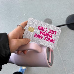 May include: A rectangular credit card skin featuring a black and white newspaper print background with the bright pink text GIRLS JUST WANNA HAVE FUNDS printed in the center, positioned above a light pink insulated tumbler with a handle.