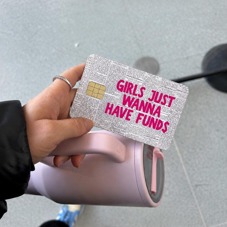 May include: A rectangular credit card skin featuring a black and white newspaper print background with the bright pink text GIRLS JUST WANNA HAVE FUNDS printed in the center, positioned above a light pink insulated tumbler with a handle.