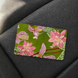 Puede incluir: Una tarjeta de cr&eacute;dito rectangular con un fondo verde oliva vibrante decorado con un patr&oacute;n repetitivo de grandes flores rosas estilo loto, follaje verde claro y peque&ntilde;as mariposas blancas, incluyendo un chip EMV dorado.