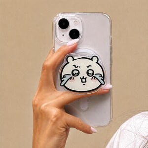 May include: A clear smartphone case featuring a circular phone grip attachment on the back. The grip displays a cartoon illustration of a white, round creature with large, teary eyes, pink cheeks, and a small mouth, outlined in bold black lines.