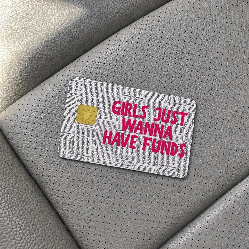 May include: A rectangular credit card skin featuring a black and white newspaper print background with the bold pink text GIRLS JUST WANNA HAVE FUNDS printed on the front. The card includes a gold-colored metallic chip on the left side.