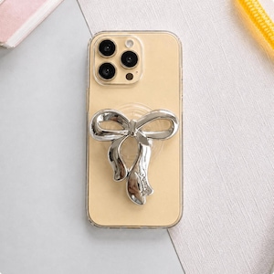 Könnte beinhalten: Eine transparente Handyhülle mit einem goldfarbenen Telefon im Inneren. Ein silberner, schleifenförmiger Handyhalter ist auf der Rückseite des Telefons angebracht. Das Telefon hat drei Kameralinsen.