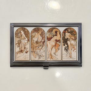 May include: A rectangular metal business card case featuring a glossy insert with four distinct vertical panels. Each panel displays a classic illustration of a figure in flowing robes, rendered in muted earth tones, cream, and soft gold hues.