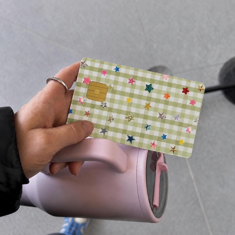 Puede incluir: Una tarjeta de cr&eacute;dito rectangular con un patr&oacute;n de cuadros vichy en verde claro y blanco, decorada con peque&ntilde;as estrellas hologr&aacute;ficas multicolores. La tarjeta incluye un chip EMV dorado en el lado izquierdo y tiene esquinas redondeadas.
