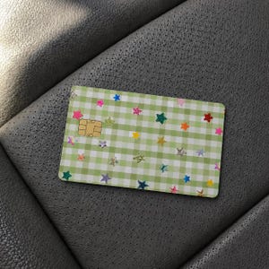 Puede incluir: Una tarjeta de pago rectangular con un patr&oacute;n de cuadros vichy en verde claro y blanco. La superficie est&aacute; decorada con numerosas estrellas peque&ntilde;as y multicolores en tonos rosa, azul, amarillo, naranja y verde. Se ve un chip met&aacute;lico dorado.