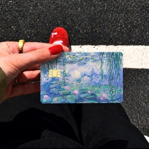 May include: A rectangular payment card featuring a printed reproduction of a water lily pond painting with shades of blue, green, and pink. The card includes a gold-colored EMV chip on the left side and displays a textured, impressionistic art style.