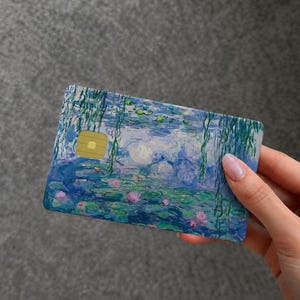 May include: A rectangular payment card featuring a printed reproduction of an impressionist water lily painting. The design displays blue and green water, pink lily pads, and hanging willow branches, with a gold metallic chip on the left side.