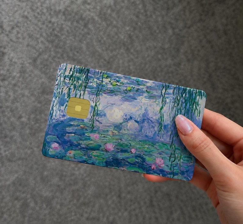 May include: A rectangular payment card featuring a printed reproduction of an impressionist water lily painting. The design displays blue and green water, pink lily pads, and hanging willow branches, with a gold metallic chip on the left side.