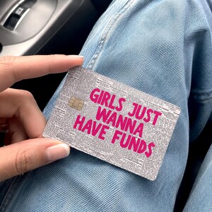 May include: A rectangular credit card skin featuring a black and white newspaper print background with the bold, bright pink text "GIRLS JUST WANNA HAVE FUNDS" printed across the center. The card includes a standard gold-colored EMV chip.