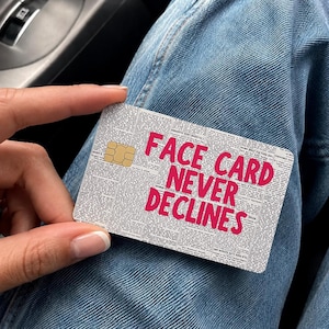 May include: A rectangular credit card skin featuring a black and white newspaper print background with the bold, bright pink text FACE CARD NEVER DECLINES printed across the center and a gold metallic chip embedded on the left side.