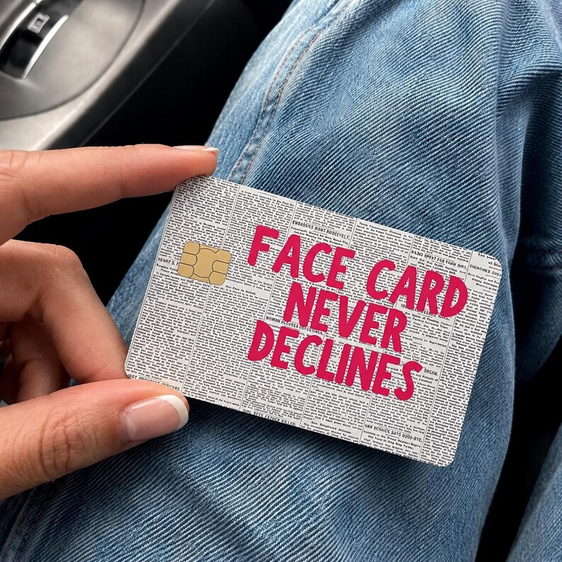 May include: A rectangular credit card skin featuring a black and white newspaper print background with the bold, bright pink text FACE CARD NEVER DECLINES printed across the center and a gold metallic chip embedded on the left side.