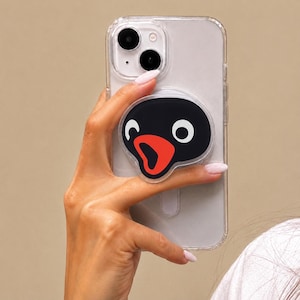 May include: A clear smartphone case featuring a circular phone grip attachment. The grip displays a graphic of a black penguin head with a large orange beak and white eyes, mimicking the character Pingu with a surprised expression.