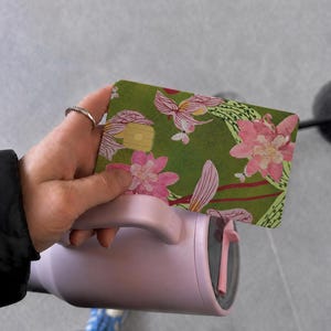 Puede incluir: Una tarjeta de pago rectangular con fondo verde y un estampado floral en rosa y blanco, sostenida sobre un vaso t&eacute;rmico de color violeta claro con asa. La tarjeta incluye un chip met&aacute;lico de color dorado en la parte frontal.