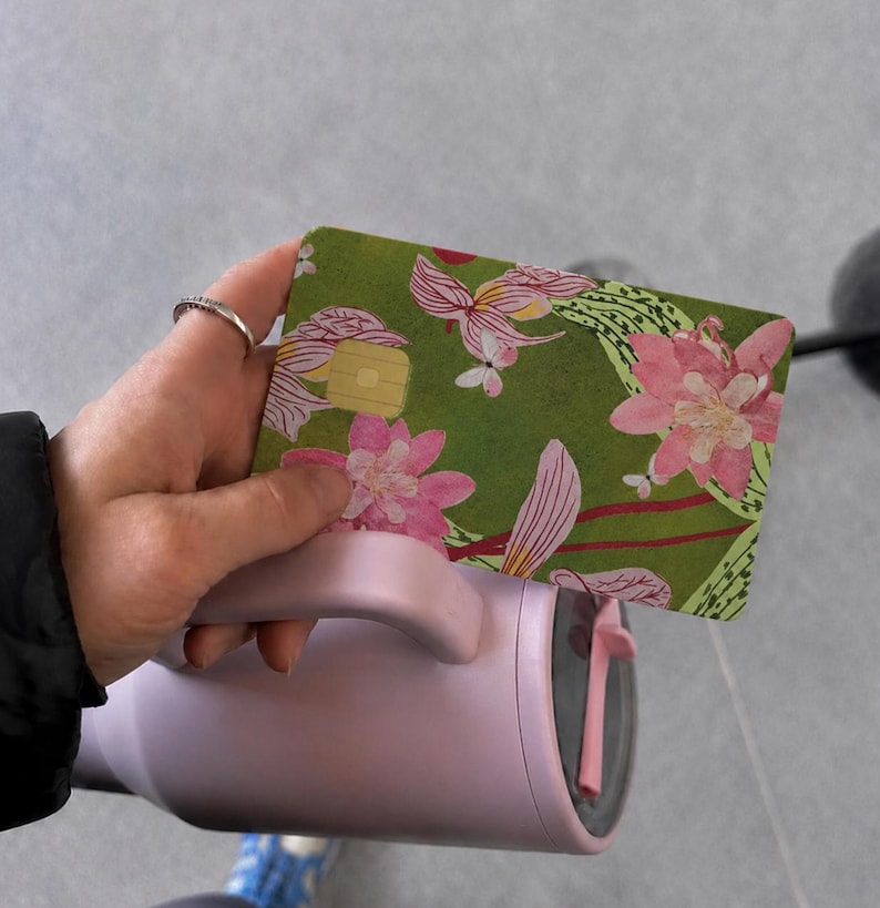 Puede incluir: Una tarjeta de pago rectangular con fondo verde y un estampado floral en rosa y blanco, sostenida sobre un vaso t&eacute;rmico de color violeta claro con asa. La tarjeta incluye un chip met&aacute;lico de color dorado en la parte frontal.