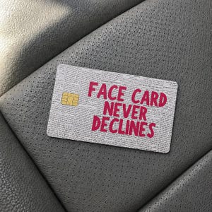 May include: A rectangular credit card skin featuring a newspaper print background with small black text and the bold, bright pink text FACE CARD NEVER DECLINES printed in the center. A gold-colored metallic chip is visible on the left side.
