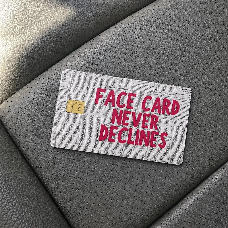 May include: A rectangular credit card skin featuring a newspaper print background with small black text and the bold, bright pink text FACE CARD NEVER DECLINES printed in the center. A gold-colored metallic chip is visible on the left side.