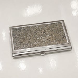 May include: A rectangular metal business card case featuring a polished silver-tone frame and a central panel with an intricate, embossed floral pattern in shades of muted gray and silver. The design displays detailed leaves and blossoms.