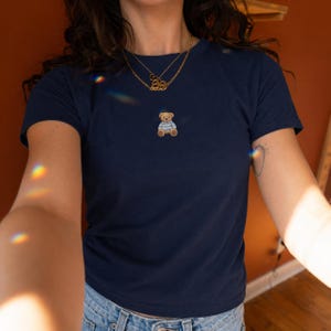 Può includere: T-shirt blu navy con un piccolo orsetto ricamato che indossa una maglietta a righe blu e bianche. La maglietta ha maniche corte e girocollo. È visibile una collana d'oro.