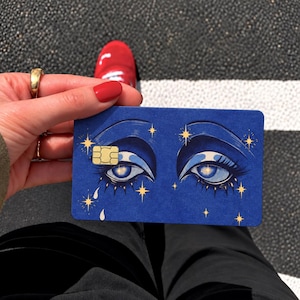 May include: A rectangular blue credit card featuring a graphic illustration of two eyes with long lashes, surrounded by small yellow star icons and white teardrop shapes. The card includes a gold-colored EMV chip on the left side.