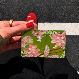 Puede incluir: Una tarjeta de cr&eacute;dito rectangular con un fondo verde vibrante decorado con un patr&oacute;n de flores rosas y blancas, hojas verdes y l&iacute;neas rojas finas. La tarjeta incluye un chip met&aacute;lico de color dorado incrustado en el lado superior izquierdo.