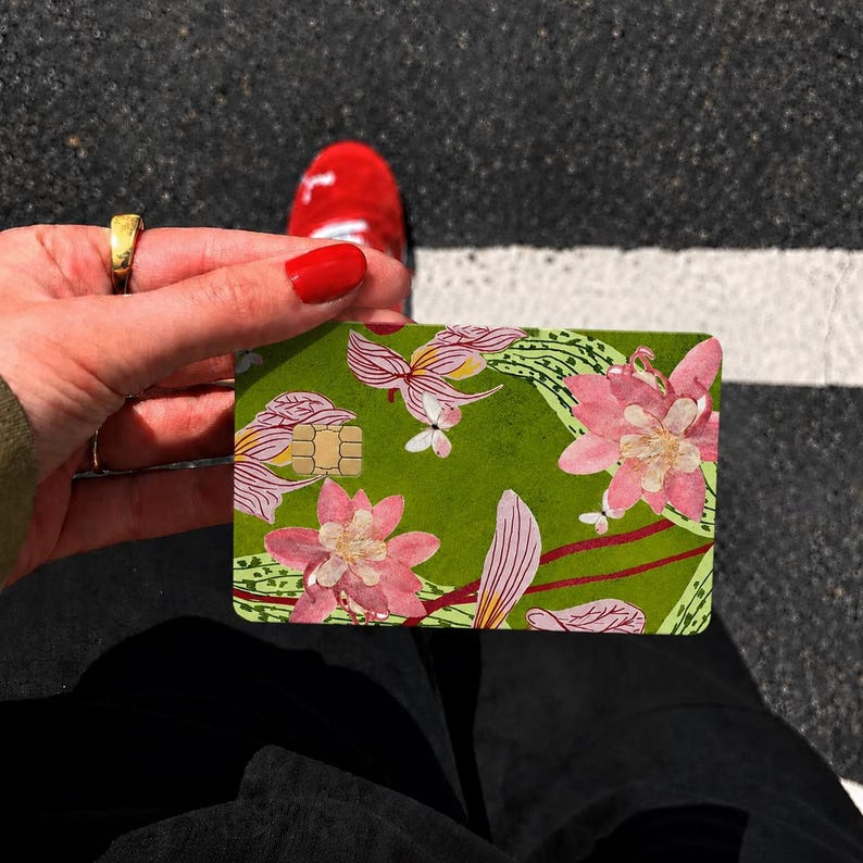 Puede incluir: Una tarjeta de cr&eacute;dito rectangular con un fondo verde vibrante decorado con un patr&oacute;n de flores rosas y blancas, hojas verdes y l&iacute;neas rojas finas. La tarjeta incluye un chip met&aacute;lico de color dorado incrustado en el lado superior izquierdo.