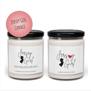 May include: Two glass candle jars with black lids. The jars are filled with a white candle and have a white label with black text that reads "Jersey Girl" and a silhouette of the state of New Jersey. The label on one jar also includes a red heart and the text "Sassy, Classy, and a Little Jersey".