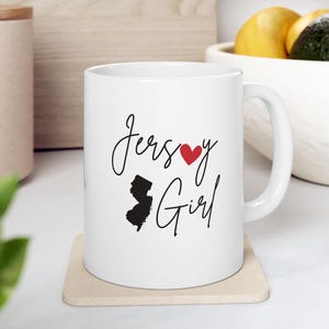 May include: White ceramic mug with a black outline of the state of New Jersey and the text "Jersey Girl" with a red heart in the word "Jersey".