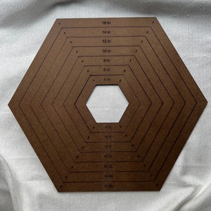 Puede incluir: Una plantilla hexagonal marrón con círculos concéntricos marcados en pulgadas de 4 a 16 pulgadas. La plantilla está hecha de cartón y tiene un agujero en el centro.
