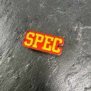 May include: A red and yellow enamel pin with the word "SPEC" in a bold, blocky font.