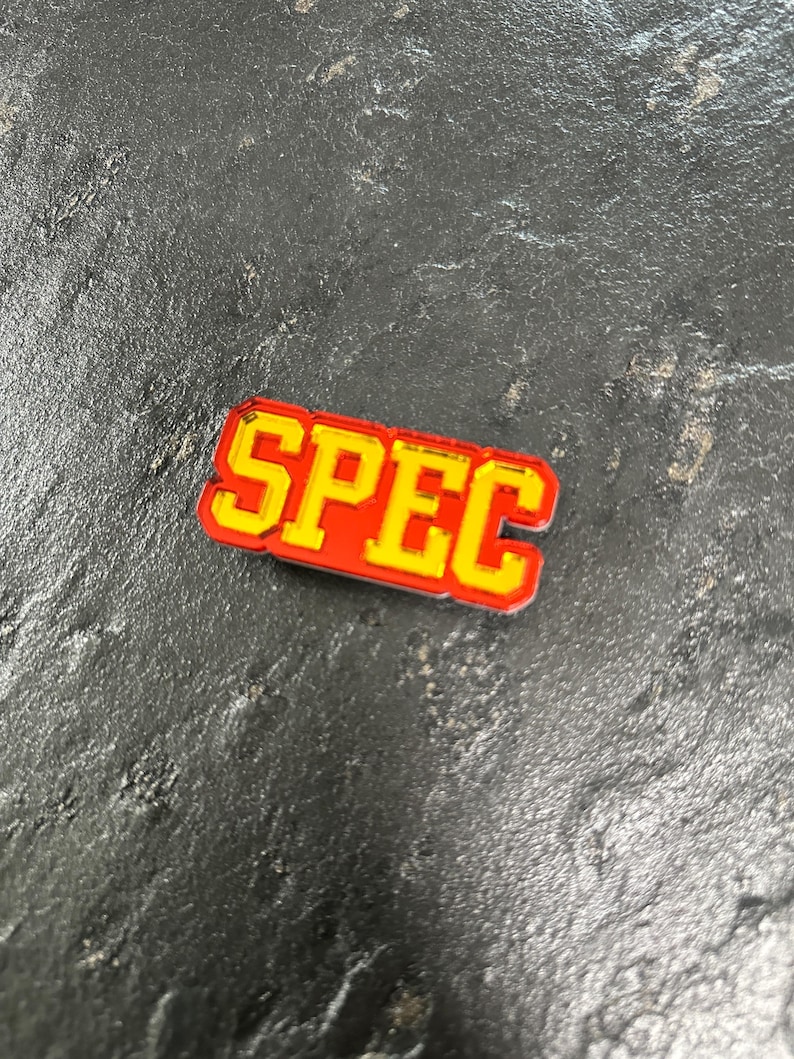 May include: A red and yellow enamel pin with the word "SPEC" in a bold, blocky font.