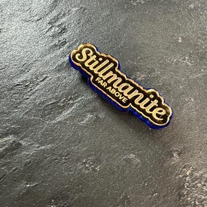 May include: A blue and gold Stillmanite pin with the words "Stillmanite" and "FAR ABOVE" in gold lettering. The pin is on a dark gray textured surface.