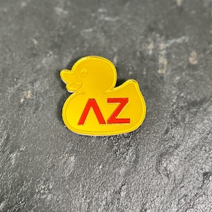 May include: A yellow rubber duck-shaped pin with the letters "AZ" in red. The pin is on a dark, textured surface. The duck has a smiling face and a white outline.