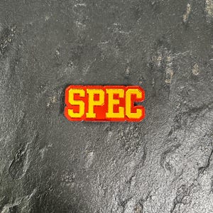 May include: A red and yellow enamel pin with the word "SPEC" in yellow letters.