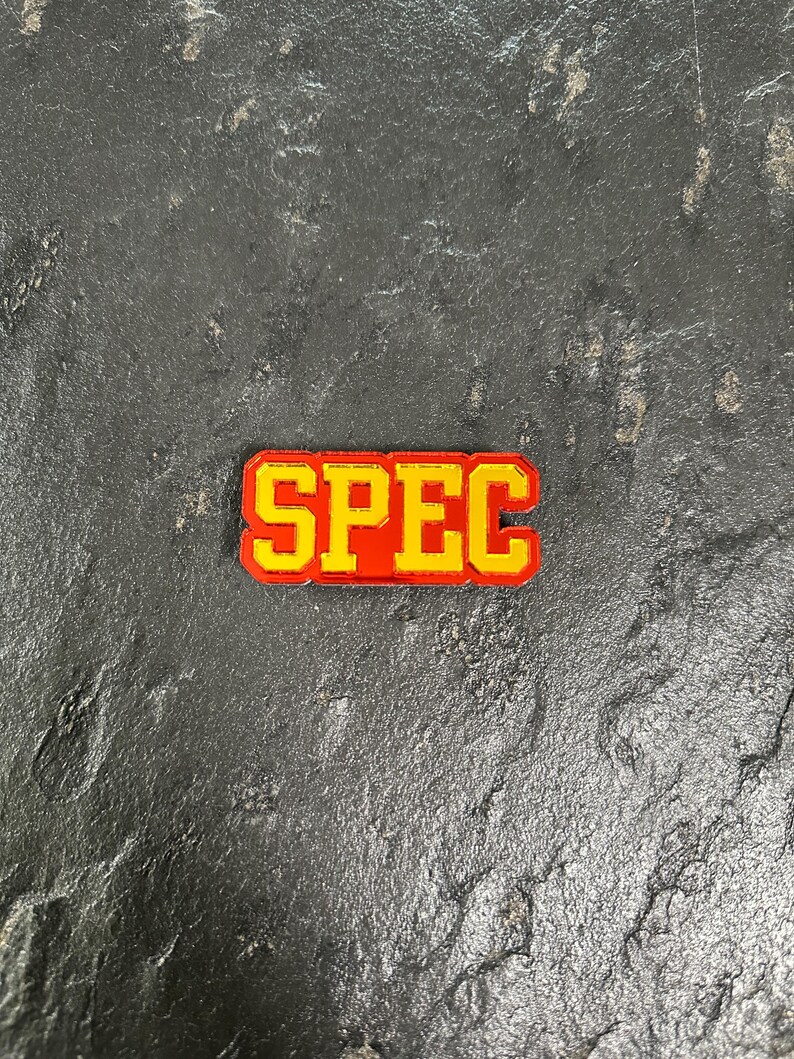 May include: A red and yellow enamel pin with the word "SPEC" in yellow letters.