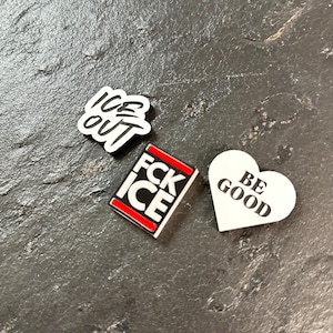 Op de afbeelding: Drie kleine, witte accessoires met zwarte tekst. Eén zegt "ICE OUT", een andere zegt "FCK ICE" in een rood omrand rechthoek, en de laatste is een hartvorm met "BE GOOD". Alle liggen op een donkere, getextureerde ondergrond.