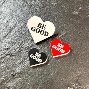 Op de afbeelding: Drie hartvormige bedels met de tekst "BE GOOD". Eén is wit met zwarte tekst, een andere zwart met witte tekst en de derde rood met witte tekst. Ze liggen op een donker, getextureerd oppervlak.