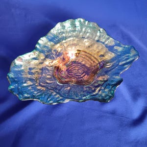 Könnte beinhalten: Eine irisierende Glasschale mit einem wellenförmigen, organischen Rand. Die Oberfläche weist ein strukturiertes, gewelltes Muster auf, das von tiefem Blau an den Rändern zu einem warmen, gold-bernsteinfarbenen Ton in der Mitte übergeht.