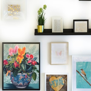 Puede incluir: Una pared de galería con arte enmarcado y elementos decorativos. Las obras de arte incluyen estampados florales, diseños abstractos de pan de oro y una ilustración de mariposa. Una pequeña estantería contiene una planta en maceta y piezas enmarcadas adicionales.