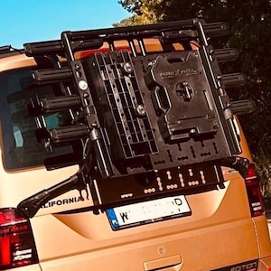 Può includere: Un furgone dorato con un portabiciclette nero sul retro. Il portabiciclette è ripiegato e ha un pannello in plastica nera con la scritta "ROTO" stampata su di esso. Il furgone ha una targa che recita "W ID".