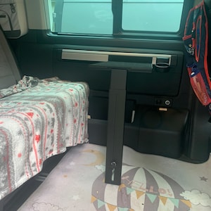 Puede incluir: Una mesa de metal negro con un portavasos en el lateral, montada en la pared de una furgoneta. La mesa está extendida y una manta gris y blanca está en el asiento al lado. El suelo está cubierto con una alfombra de color gris claro y blanco con un diseño de globo aerostático y jirafa.
