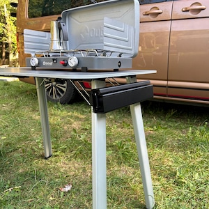 May include: A portable camping stove with a black and silver design. The stove has two burners and a wind shield. The stove is sitting on a table with a black and silver design. The table has four legs and is made of metal. The stove is next to a brown van.