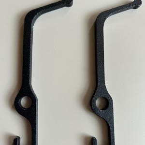 Pode incluir: Dois ganchos de metal preto com a parte superior arredondada e um gancho na parte inferior. Os ganchos são projetados para pendurar objetos.