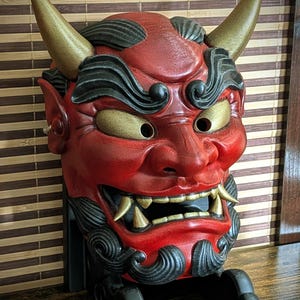 Könnte beinhalten: Eine rote, schwarze und goldene japanische Dämonenmaske mit Hörnern und einem grimmigen Gesichtsausdruck. Die Maske hat goldfarbene Hörner, Augen und Zähne sowie schwarze Haar- und Bartdetails. Die Maske ist auf einem schwarzen Ständer montiert.