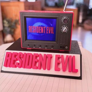 May include: A small red and black television model, with the words "RESIDENT EVIL" visible on the screen and base. The screen displays a blue background with the same text in red. The TV has dials and an aerial.