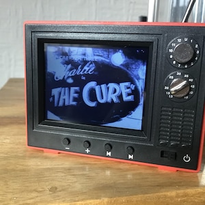 Può includere: Un televisore retrò rosso e nero con uno schermo che mostra un'immagine in bianco e nero della band "The Cure". Il televisore ha un quadrante con i numeri da 13 a 40 e un pulsante di accensione.