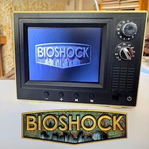 Könnte beinhalten: Ein kleiner, gelb-schwarzer Fernseher mit dem Wort "BIOSHOCK" auf dem Bildschirm. Darunter befindet sich ein passendes Schild mit dem Logo des Spiels. Der Fernseher hat Knöpfe und Drehregler.